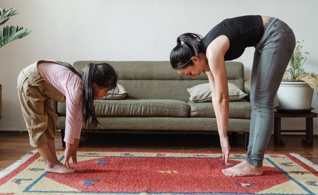 Side view of flexible young ethnic mother exercising with cute daughter while standing against each other on floor carpet near cozy sofa in modern living room in morning